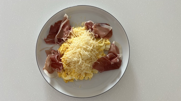 Scrambled eggs topped with shredded cheese and slices of prosciutto